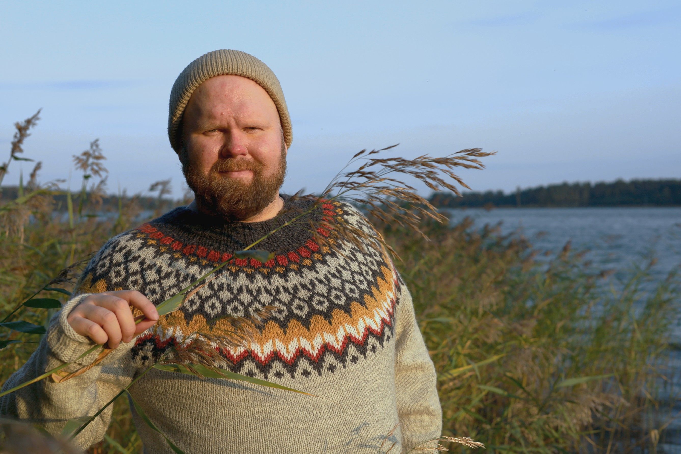 Antti Brunni in a wool sweater and beanie among lakeside reeds, holding a reed stem.