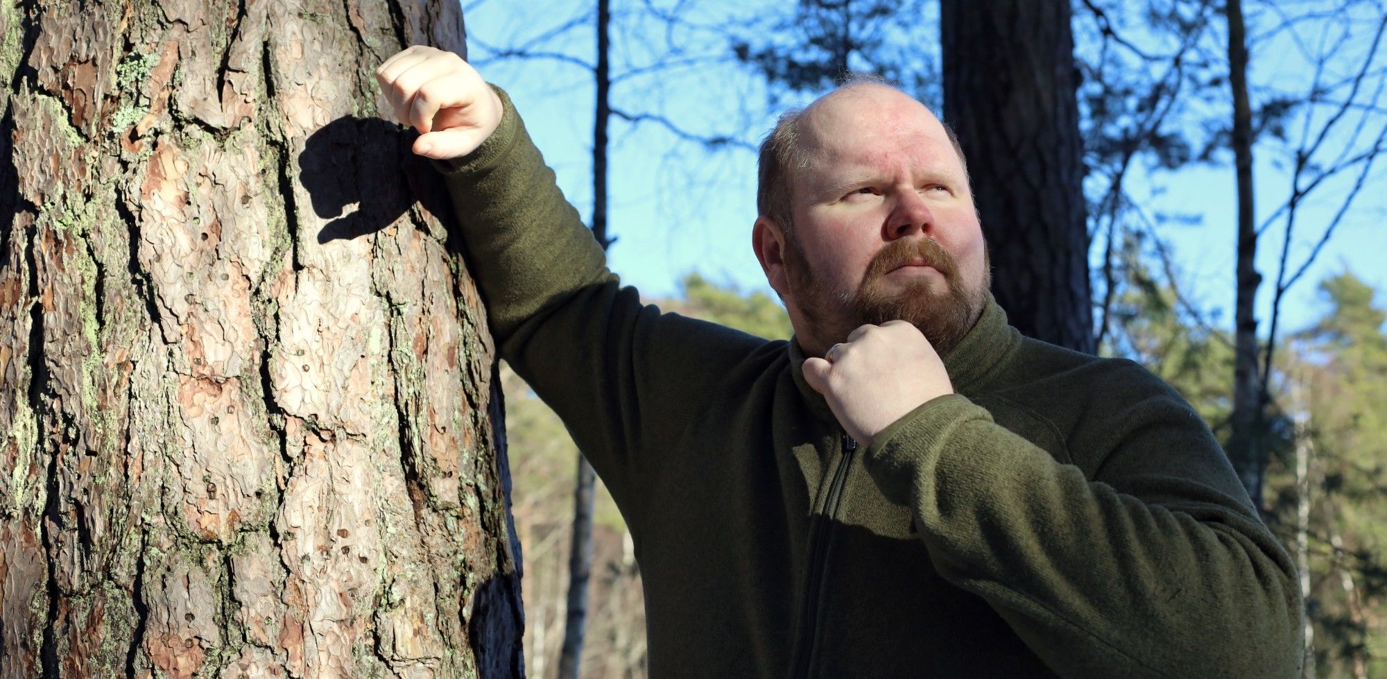 Antti Brunni leaning against a pine trunk in a sunlit forest, looking thoughtfully to the side.